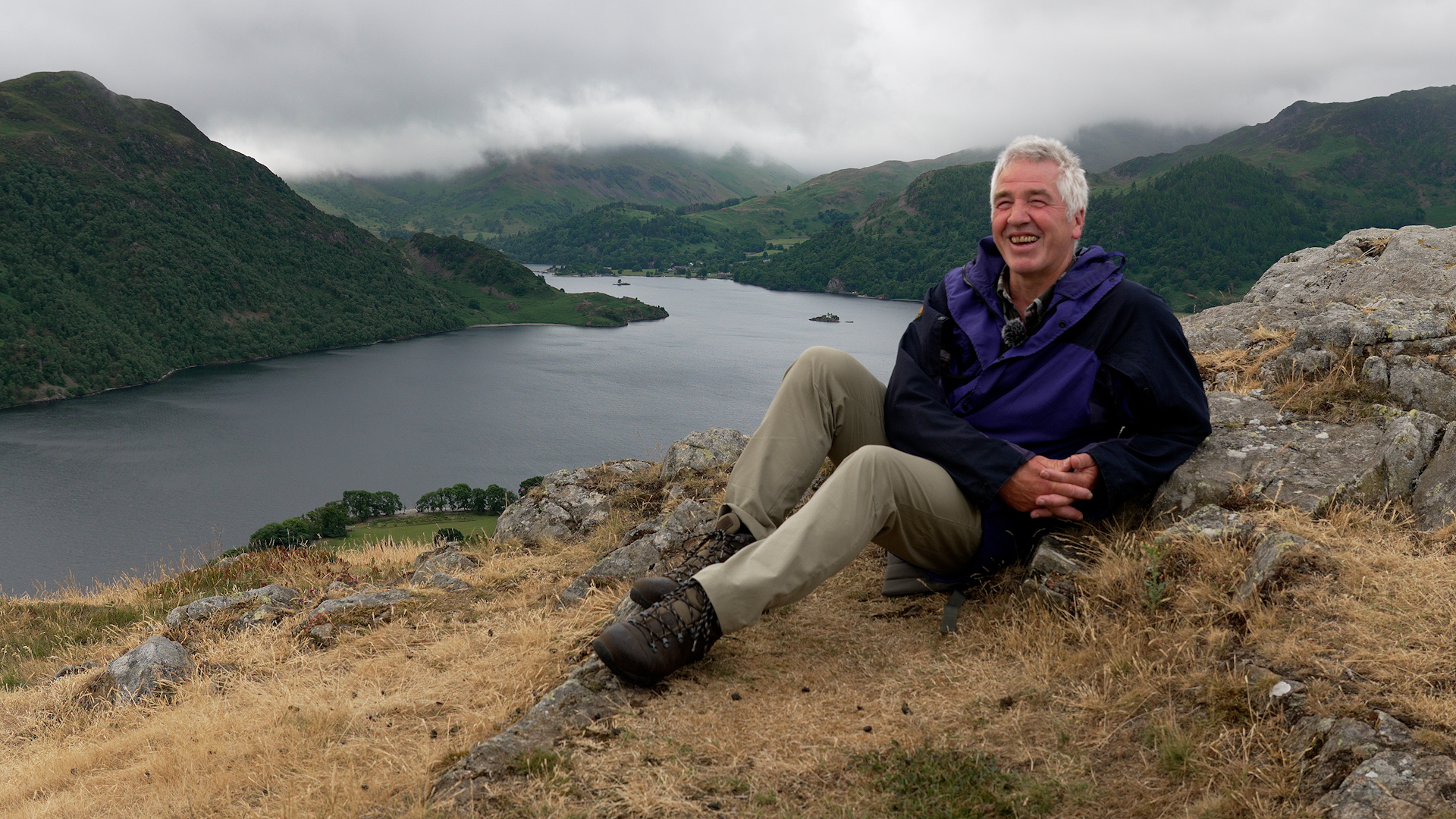 A man sits on a rocky outcrop, with a lake and hills behind him