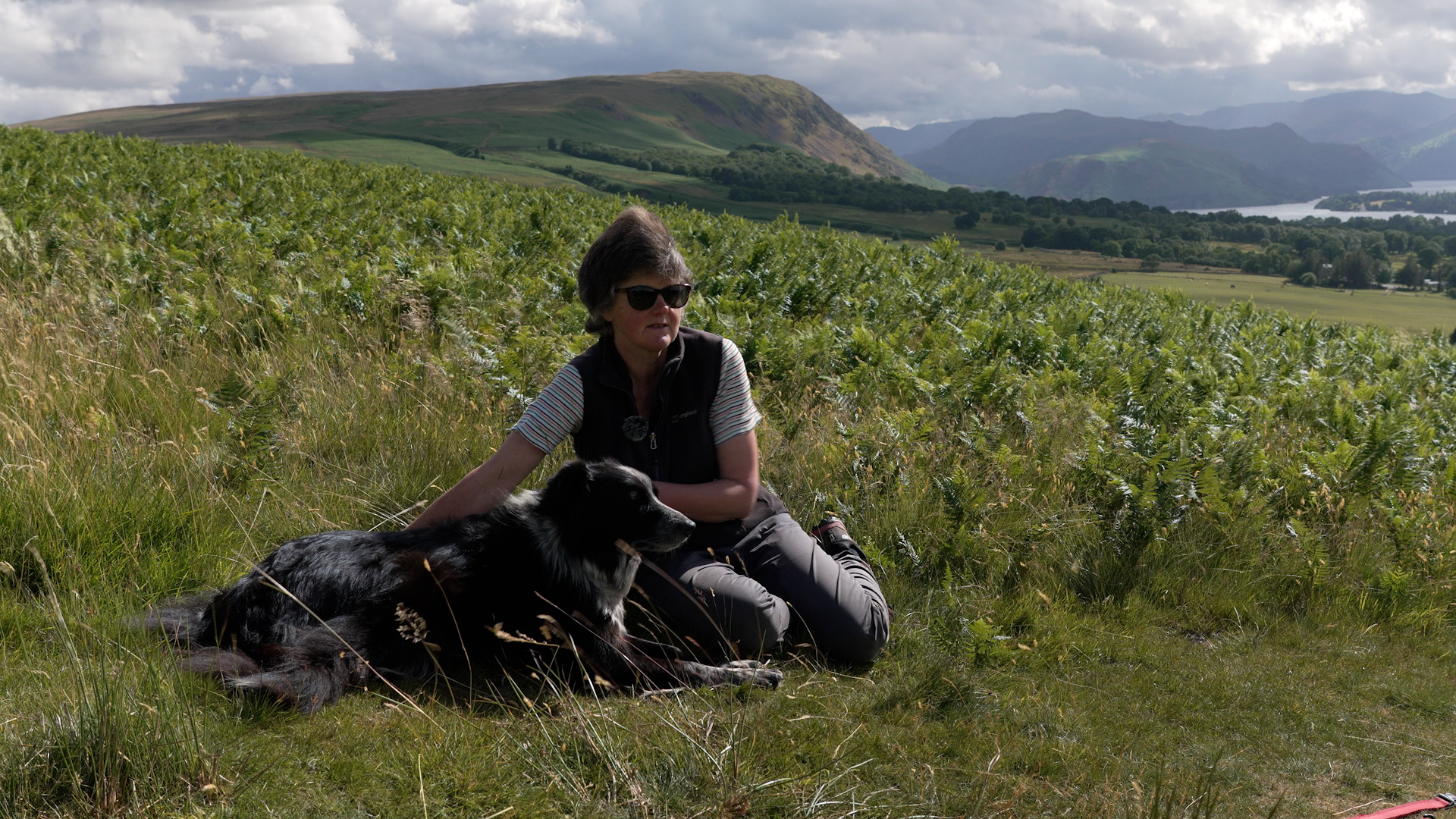 A woman sits with her dog on a grassy hillside, with hills and a lake in the distance