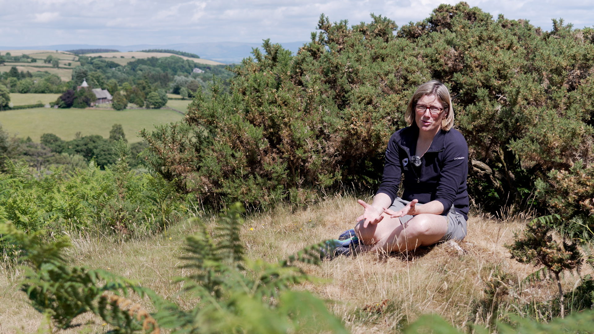 A woman sits on a patch of parched grass with a gorse bush behind her