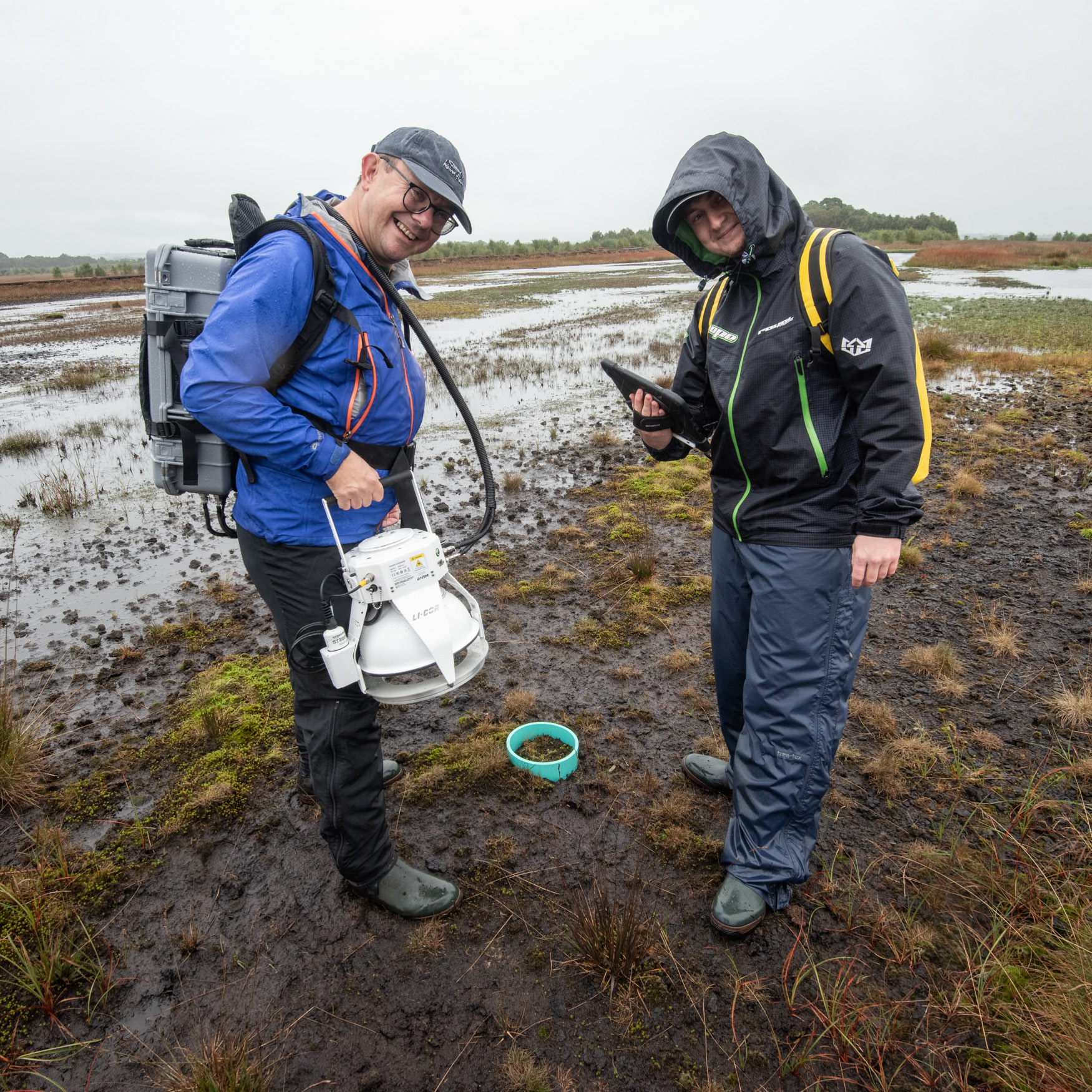 Two men in raincoats stand on a wet bog and look at the camera; the man on the left holds a white object, which is a monitor he will use to measure carbon dioxide and methane emissions