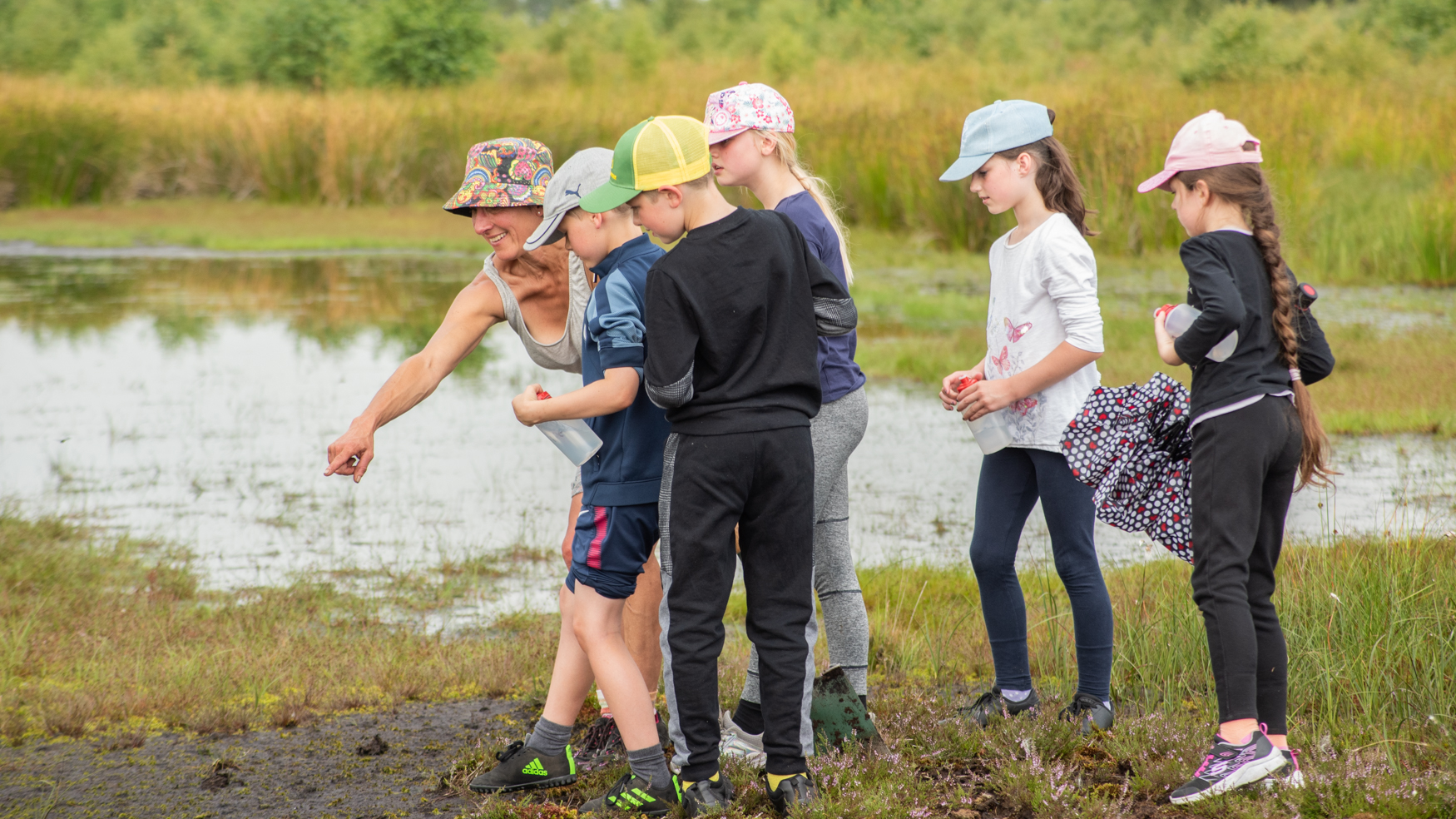 A woman in a colourful hat points at something on the ground. Five children, wearing caps, look where she is pointing. They are standing in a boggy area with a pond behind them, and long grasses.
