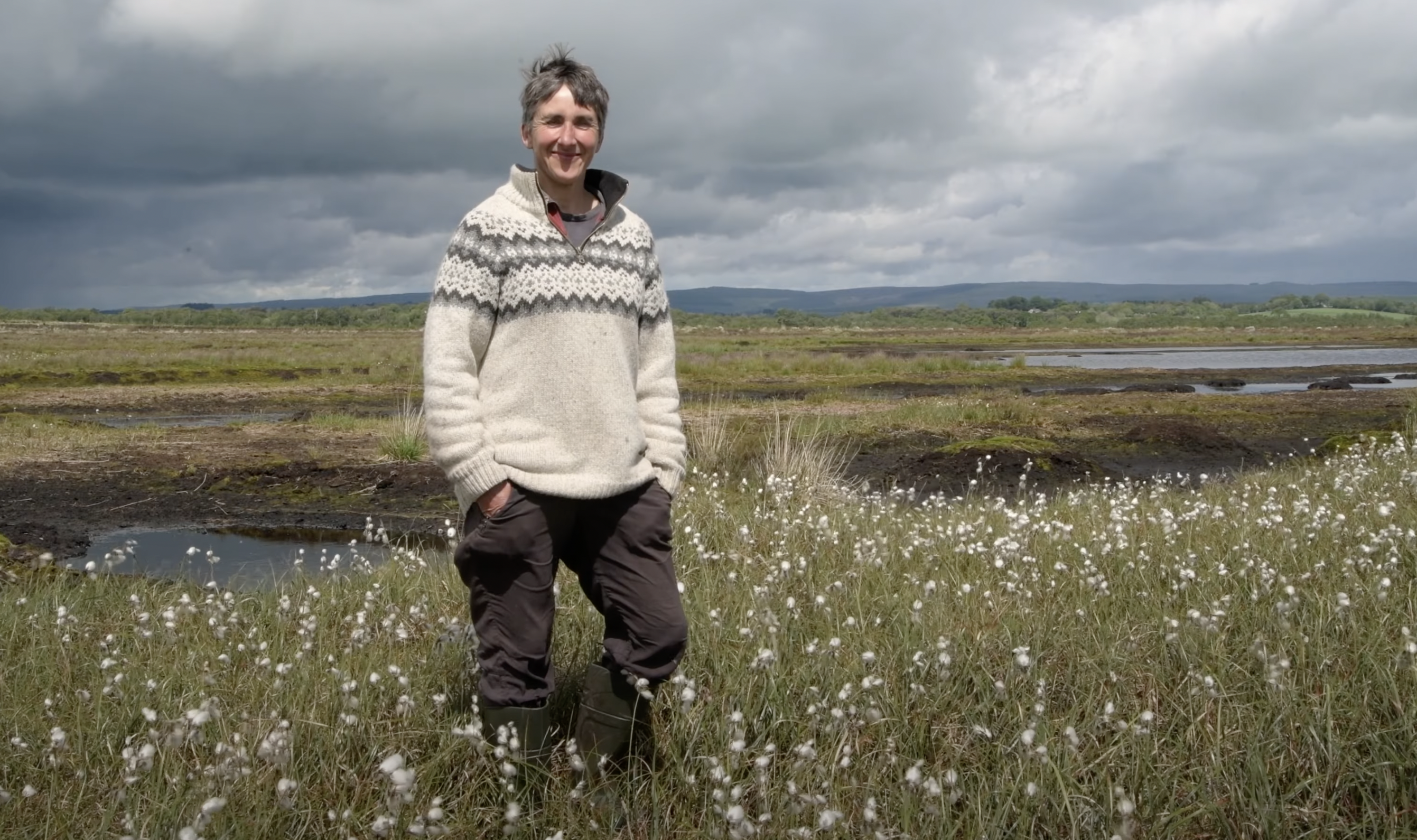 A woman in a pale jumper stands among cotton grass, with a grey cloudy sky