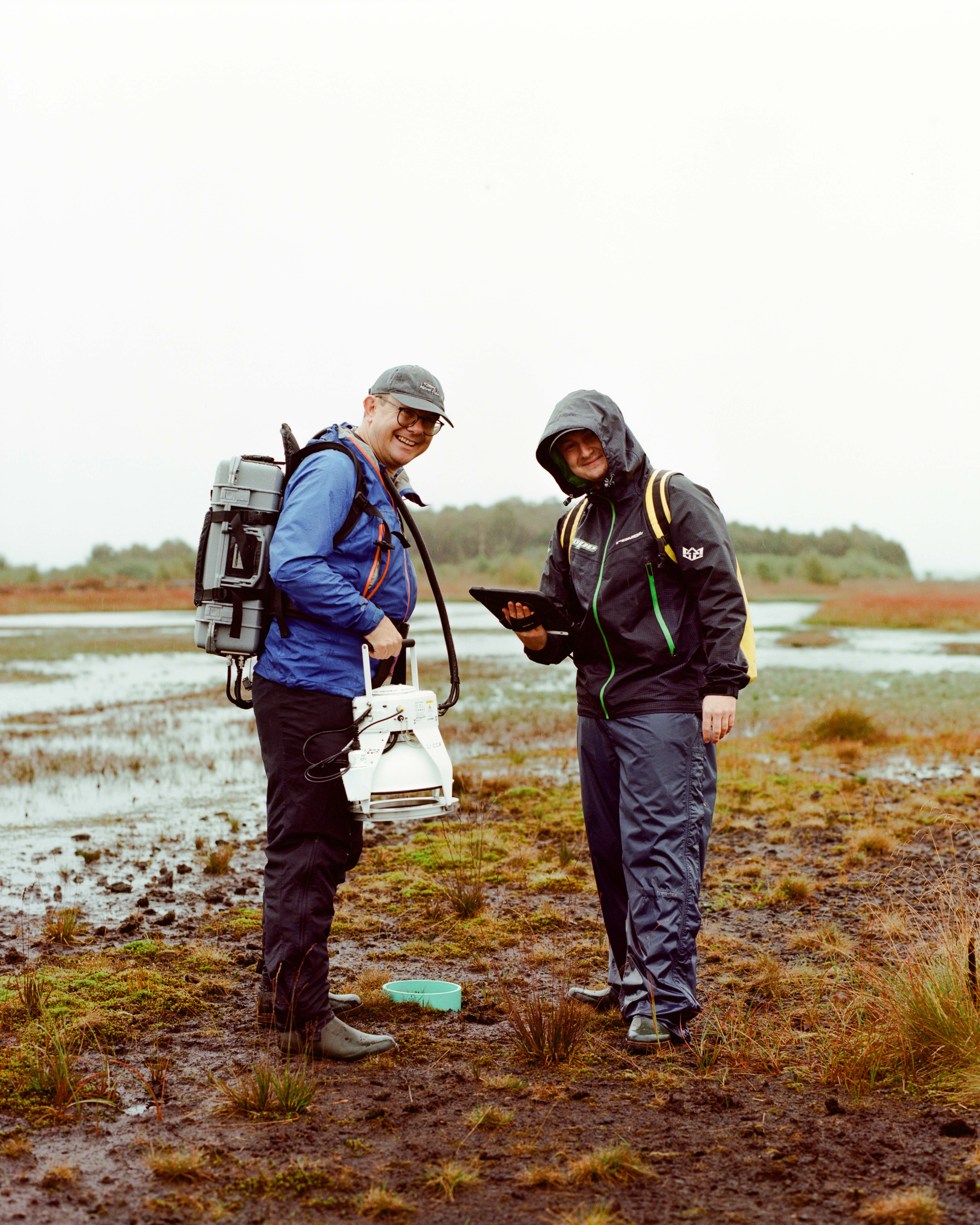 Two men standing on a bog look towards the camera; they are dressed in waterproofs.